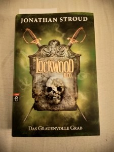 Titebild des Buches "Lockwood & Co. - Das Grauenvolle Grab" von Jonathan Stroud Ein Grabstein mit Totenschädel, auf dem Lockwood & Co. steht, und hinter dem sich zwei Rapiere kreuzen Hinter dem Grabstein scheint es zu glühen.