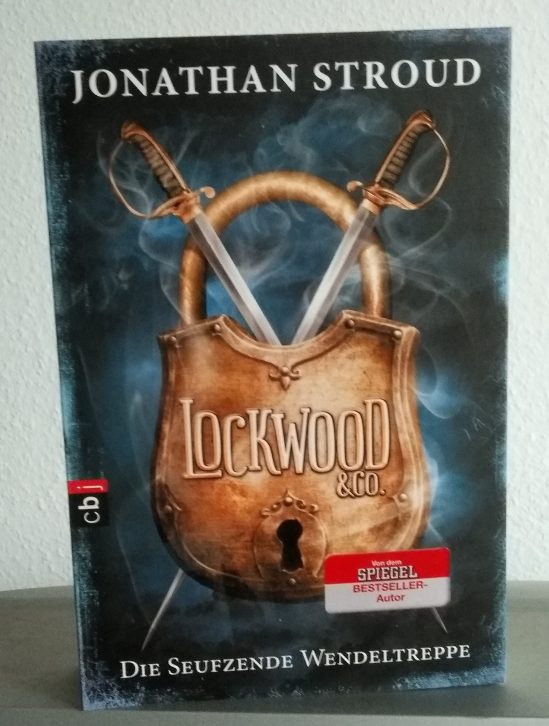 Titelbild des Buches "Lockwood & Co. - Die seufzende Wendeltreppe" von Jonathan Stroud
Ein großes altes Vorhängeschloss, auf dem Lockwood & Co. steht, und hinter dem sich zwei Rapiere kreuzen