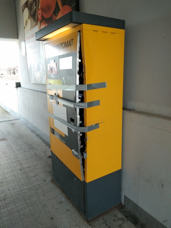 Parkautomat, dessen Vorderseite nur noch mit Klebestreifen festgehalten wird und mit einem Spalt absteht. Wahrscheinlich aufgebrochen oder gar gesprengt.