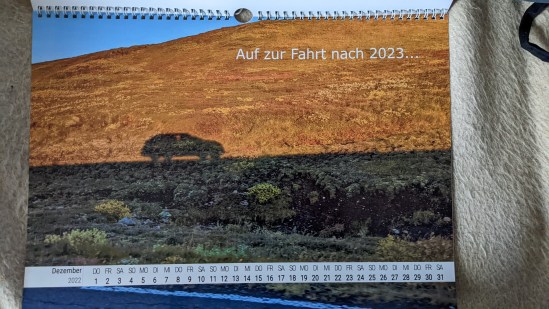 Dezember Der Schatten eines Autos auf einem grün braunen Feld. Rechts oben steht: Auf zur Fahrt nach 2023...