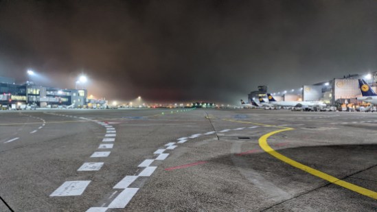Leeres Vorfeld im Dunkeln, hell erleuchtet durch die Scheinwerfer. Auf der rechten Seite stehem ein paar Flugzeuge an den Gates. Der Dunst ist in leichten Schwaden am Himmel zu sehen.
