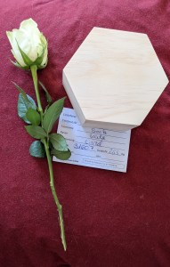 Auf rotem Hintergrund liegt eine weiße Rose, ein sechseckiges Holzkästchen und daruter ein Zettel, auf dem zu lesen ist: Einlieferer Tierbesitzer Tiername: Socke Rasse: Katze ...mierung: einzel Nr. 31607 Gewicht: 2,55 kg (Material der Kategorie 1 nur zur Entsorgung)