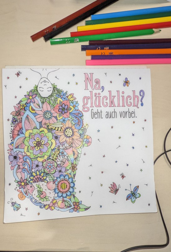 Ausmalbild: Oben ein Frauenkopf, der nach unten eine Art Frisur aus lauter Blumen und Blättern hat, zusammen mit ein paar Schmetterlingen, Käfer und Biene Dazu steht neben dem Kopf dieser Text: Na, glücklich? Geht auch vorbei. Über dem Ausmalbild liegen noch ein paar Buntstifte.
