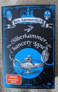 Buchtitel Ben Aaronovitch "Die Silberkammer in der Chancery Lange" In einem blauen Kreis auf schwarzem Hintergrund steht eine weiße Öllampe mit totenköpfiger Deckelfigur, aus deren Dochtöffnung etwas wie weiße Flammen entweicht.