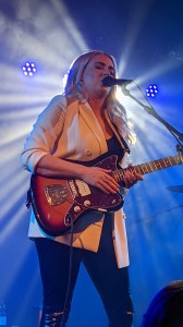 Eivør in hellem Blazer und Gitarre in den Händen, im Gegenlicht stehend gerade ins Mikrofon singend.