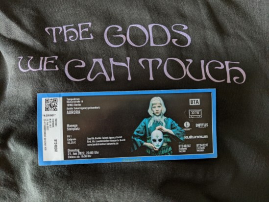 Eintrittskarte zu Konzert von Aurora im Tempodrom Berlin am 21. Juni 2022, 20 Uhr - Manege Stehplatz Aurora in ihrem grünen Kleid halt die bemalte weiße Maske vor sich. Die Karte liegt auf einem dunklem Hintergrund, auf dem steht: THE GODS WE CAN TOUCH