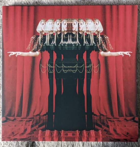 Cover des Albums "The Gods We Can Touch" von Aurora Aurora in einem schwarzen Kleid, die Arme ausgebreitet, Prismenartig zu sehen, vor einem roten Vorhang.