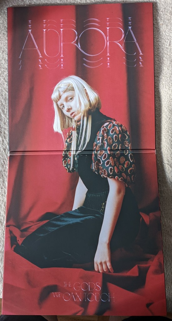 Innenseite des Klappcovers Aurora in ihrem schwarzen Kleid, auf und vor einem roten Tuch und Vorhang knieend, Format hochkant Unten steht THE GODS WE CAN TOUCH