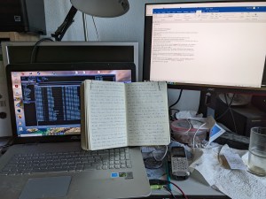 Auf dem Schreibtisch steht links ein Laptop vor einem Monitor, auf dem das aufgeklappte Notizbuch steht. Rechts ist der Monitor mit der geöffneten Textdatei.