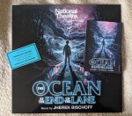 Cover des Soundtracks zum Theaterstück des National Theatre "The Ocean at the End of the Lane" Music from Jherek Bischoff Ein Junge steht mit dem Rücken zum Betrachter im Gegenlicht auf einem Feldweg, links und rechts steht das Wasser. Links ist ein dunkler Wald , vor dem Jungen türmen sich Wolkenberge. Im Zentrum der Wolkenberge über dem Junge sind Blitze zu sehen. Rechts auf dem Cover liegt noch eine Postkarte mit dem gleichem Motiv.