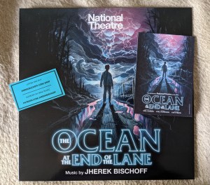 Cover des Soundtracks zum Theaterstück des National Theatre "The Ocean at the End of the Lane" Music from Jherek Bischoff Ein Junge steht mit dem Rücken zum Betrachter im Gegenlicht auf einem Feldweg, links und rechts steht das Wasser. Links ist ein dunkler Wald , vor dem Jungen türmen sich Wolkenberge. Im Zentrum der Wolkenberge über dem Junge sind Blitze zu sehen. Rechts auf dem Cover liegt noch eine Postkarte mit dem gleichem Motiv.