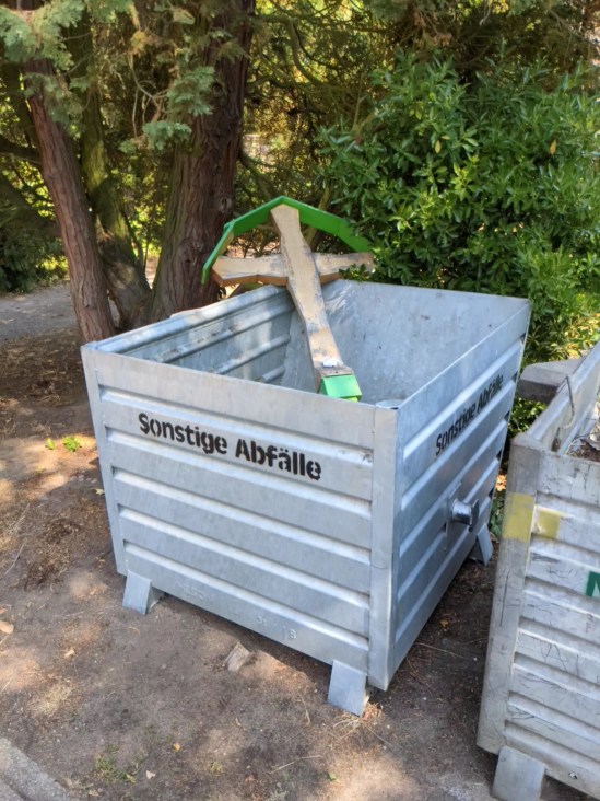 In einem Abfallbehälter mit der Aufschrift "Sonstige Abfälle" liegt ein hölzernes Grabkreuz ohne Beschriftung
