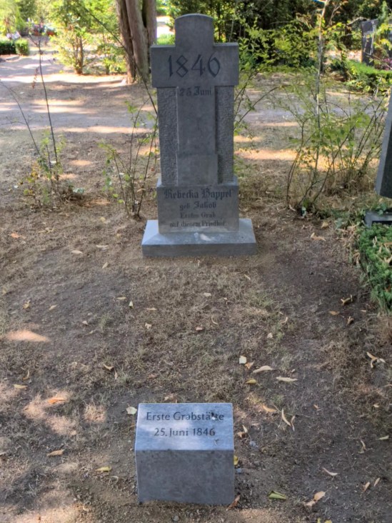 Grabstein mit Kreuz auf dem steht: Rebecka Kappel, geb. Jakob Erstes Grab auf diesem Friedhof, davor ein Gedenkstein: Erste Grabstätte 25. Juni 1846