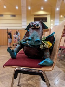 Handpuppen-Drache in braun, mit hellblauen Pfoten, hellblauer Schnauze und hellblauen Rändern an den Flügeln mit einer gelben Rose im Maul, auf einem Stuhl sitzend