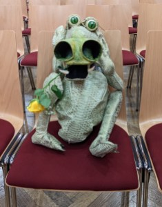Handpuppendrache Eroll auf einem Stuhl, mit gelber Rose im Maul. Seine Farbe ist grünlich - und er hat zwei extra große Nasenlöcher