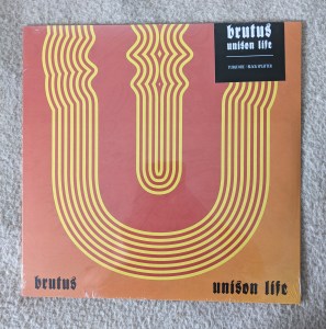 LP-Cover der Band brutus - unison life Es ist ein stilisiertes U zu erkennen, dargestellt durch gelbe Streifen vor einem rot bis orangenem Hintergrund