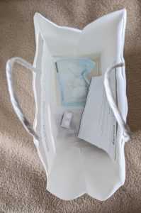 Weiße Care-Papiertüte, in der verpackte Tupfer-Kompressen, zwei Schmerztabletten, ein kleines Kühlpaket und ein Schreiben mit Hilfestellungen befindet