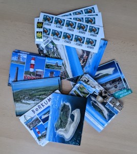 8 verschiedene Ansichtskarten von Borkum rund aufgefächert. Es ist zu erkennen, dass es mehrere Karten von den einzelnen Varianten gibt. Oben liegen 4 Zehnerstreifen der Briefmarke für 70 ct. Außerdem 2 Briefmarken zu 10 ct und eine zu 5 ct.