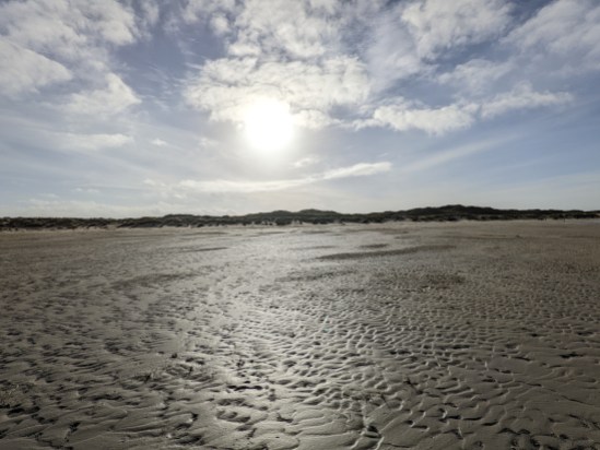 Bis zur Hälfte des Bildes ist der furchige Sandstrand zu sehen. In der Mitte des Bildes ist die Dünenlinie zu erkennen. Darüber scheint die Sonne von einem blauen, wolkigem Himmel