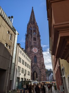 Turm mit Uhr des Freiburger Münsters, zu sehen aus einer Häusergasse heraus Vor dem Münster und in der Gasse sind Marktstände aufgebaut