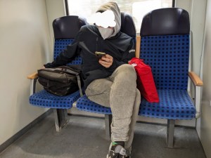 3er-Sitzgruppe in der Regionalbahn Ein Typ sitzt auf dem mittleren Platz, sein Rucksack rechts neben sich liegend auf dem Platz, auf der anderen Seite eine weitere Tasche, die auch den dritten Platz blockiert