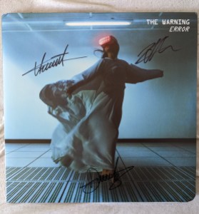 Das Cover der LP "ERROR" von The Warning, mit den Autogrammen der Band. Vor dem Hintergrund eines nüchternen Raumes mit Fußbodenplatten und Neonröhre steht eine Frau in einem wallendem Gewand, mit einer rötlich leuchtenden VR-Brille vor den Augen.