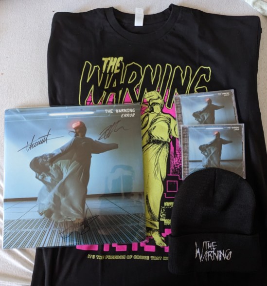 Merchandise der Band "The Warning" Eine Vinyl-Platte von ERROR mit Autogrammen; eine schwarze Mütze mit dem Schriftzug "The Warning"; zwei CDs des Albums ERROR und hinter/unter allem ein T-Shirt mit dem Motiv des Covers von ERROR - eine Frau in wallendem Gewand mit VR-Brille