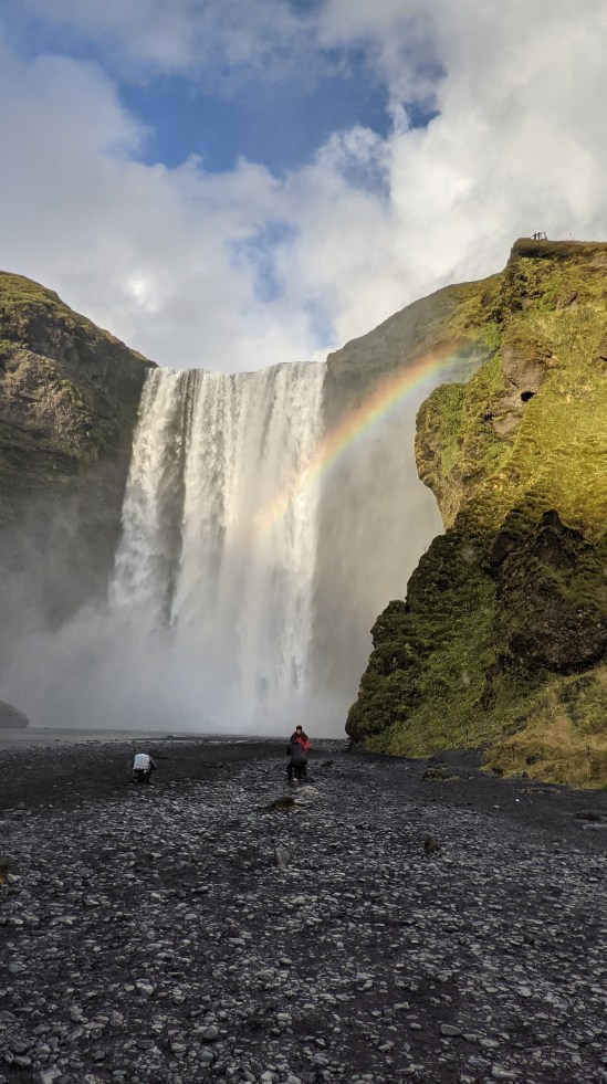 Der Wasserfall Skógafoss mit Regenbogen Vor dem Wasserfall sind auf dem Geröll drei Leute zu sehen.