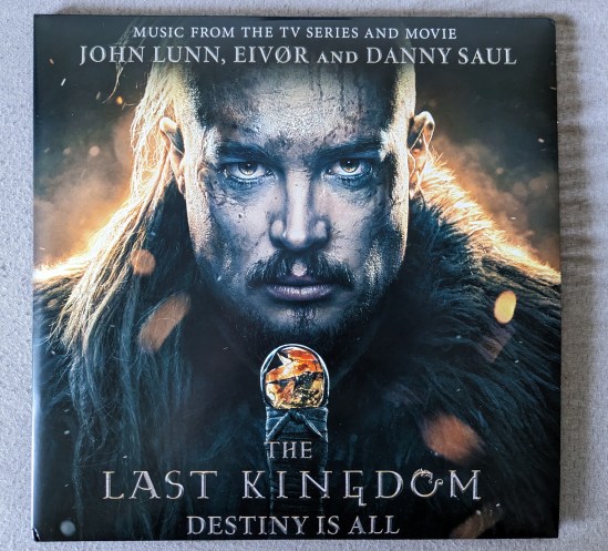 Cover der Doppel-LP THE LAST KINGDOM Destiny is all Music from the TV Series and Movie John Lunn, Eivør & Danny Saul Zu sehen ist das bärtige Gesicht eines Mannes, der in Fell gekleidet zu sein scheint und den Griff eines Schwertes vor sich hält. Von dem Schwert ist wirklich nur die oberste Hälfte des Griffes zu sehen, mit dem Knauf, der aus Bernstein zu bestehen scheint. Hinter dem Mann ist es hell, das Fell am Rand leuchtet und es sind herumfliegende Funken zu sehen.