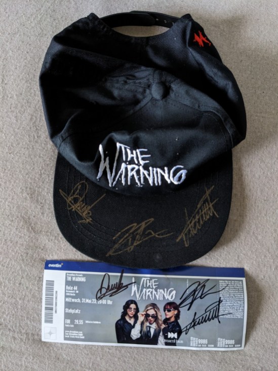 Eintrittskarte zum Konzert von The Warning in Berlin am 31. Mai 2023 und schwarze Kappe mit Aufschrift "The Warning" - auf beiden sind die Autogramme von Dany, Pau und Ale
