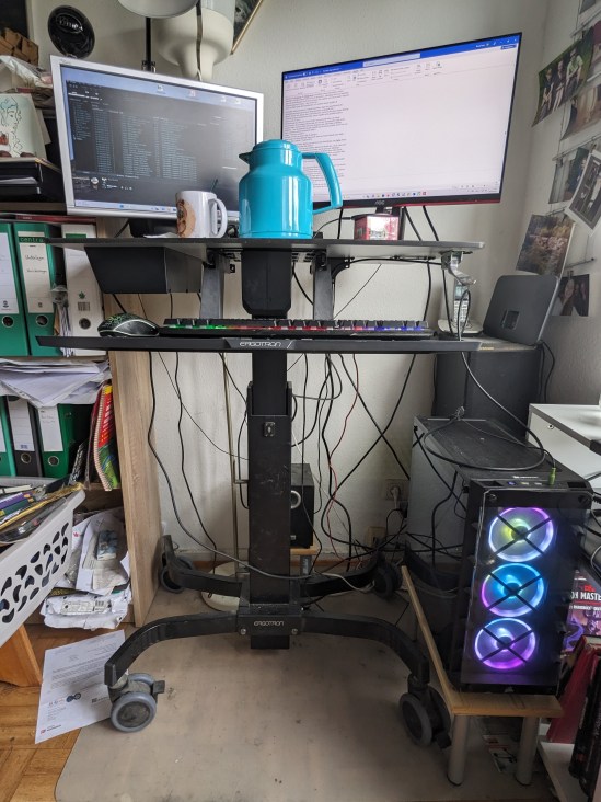 Hochgefahrener Schreibtisch mit Teekanne und Tasse vor den zwei Monitoren. Unten hinten ist der Subwoofer zu sehen, rechts steht der große Rechner, das Monster.