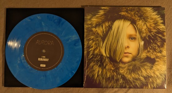 Vinyl-Platte von AURORA RUNAWAY in blauem Vinyl Daneben das Cover mit dem Portrait von Auroea, umgeben von ihrer Pelzkapuze