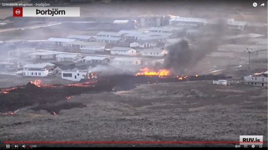Bildschirmfoto der Eruption vom 14.01.2024 Livestream von RÚV. Oben links im Eck steht die Standortangabe der Kamera: Þorbjörn Zu sehen sind einige weiße Häuser von Grindavík. Vor ihnen zieht sich ein schwarzer Lavastreifen, in dem an einigen Stellen auch noch rote Lava zu sehen ist, von links nach rechts ins Bild. Ein Haus links neben der Lava brennt , und im Bildmittelpunkt sind nur noch die Flammen eines Hauses zu sehen, welches dort steht/stand. Unten rechts steht RUV.is