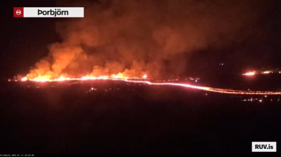 Bildschirmfoto der Eruption vom 14.01.2024 Livestream von RÚV. Unten links steht in kleiner Schrift Grindavík 2024-01-14 18:34:38 Oben links im Eck steht die Standortangabe der Kamera: Þorbjörn Das Bild ist wieder dunkel, es ist wieder Nacht, und quer durch das Bild ziehen sichdie Flammen und der glühende Streifen des Lavastroms des momentanen Ausbruchs. Im Hintergrund rechts sind ebenfalls Flammen zu sehen, wahrscheinlich durch die zweite Spalte und den brennenden Häusern. Unten rechts steht RUV.is