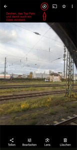 Screenschot eines Fotos, auf denen Bahngleise und ein Regenbogen zu sehen ist Über dem Foto ist von links nach rechts zu sehen: Der Pfeil nach links zum Zurückgehen, ein Dreieck in einem geöffneten Kreis zum Abspielen des Videos, das Zeichen zum Streamen, ein Stern und dre senkrechte Punkte. Unter dem Foto sind die Funktionen für Teilen, Bearbeiten, Lens und Löschen. Das Dreieck ist mit einem roten Kreis markiert und einem roten Pfeil, der darauf zeigt. Daneben steht: Zeichen, das Top-Foto und damit ein Video existiert.