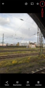 Screenschot eines Fotos, auf denen Bahngleise und ein Regenbogen zu sehen ist Über dem Foto ist von links nach rechts zu sehen: Der Pfeil nach links zum Zurückgehen, ein Dreieck in einem geöffneten Kreis zum Abspielen des Videos, das Zeichen zum Streamen, ein Stern und dre senkrechte Punkte. Unter dem Foto sind die Funktionen für Teilen, Bearbeiten, Lens und Löschen. Die drei Punkte sind mit einem roten Kreis und einem roten Pfeil markiert.