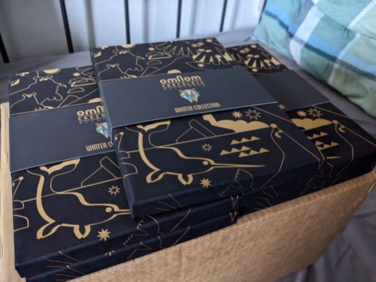 5 Packungen der OMNOM Chocholate WINTER COLLECTION. Eine schwarze Verpackung mit der Zeichnung eines Narwals und anderer Tiere in Gold,
