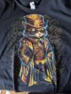 T-Shirtmotiv: Illustration einer Katze im Steampunk-Outfit mit Mantel, Zylinder, Weste und Googles in Gold gezeichnet auf schwarzem Hintergrund.