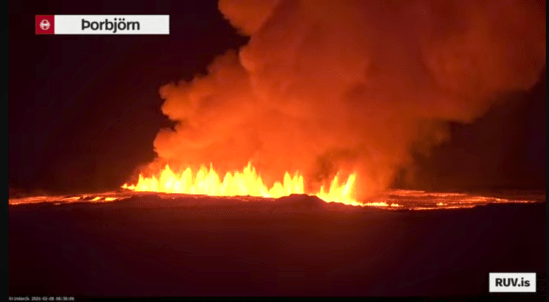 Bildschirmfoto der Eruption vom 08.02.2024 Livestream von RÚV. Unten links steht in kleiner Schrift Grindavík 2024-02-08 06:36:06 Oben links im Eck steht die Standortangabe der Kamera: Þorbjörn Es ist Nacht, der Himmel und der Vordergrund ist dunkel. In der Mitte des Bildes ziehen sich die Feuerfontänen des aktuellen Ausbruchs durch die Hälfte des Bildes. Links und rechts davon ist glühende Lava zu erkennen und eine große Rauchwolke erhebt sich über dem Eruptionsstreifen. Unten rechts steht RUV.is