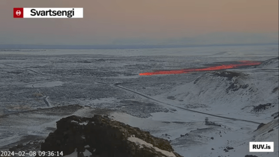 Bildschirmfoto der Eruption vom 08.02.2024 Livestream von RÚV. Unten links steht 2024-02-08 09:36:14 Oben links im Eck steht die Standortangabe der Kamera: Svartsengi Ein kühles Morgengrauen liegt über einer schneebedeckten flachen Landschaft. Durch das Bild zieht sich von rechts unten nach links Mitte eine Straße. In der Mitte des Bildes rechts, hinter ein paar Hügeln, zieht sich auf dem schneebedeckten untergrund eine rot glühende Lavazunge nach links, Richtung Bildmitte und der Straße zu. Unten rechts steht RUV.is