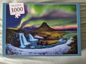 Puzzlebox von einem 1000-Teile Puzzle Das Motiv ist DAS typische Motiv des Snaefellsnes National Park: Der Berg Kirkjufell im Bildmittelpunkt hinten, davor der Kirkjufellsfoss-Wasserfall mit Eisschicht davor - und darüber Polarlichter