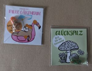 Zwei ein kleinen Tüten verpackte Samenpäckchen. Eines nennt sich Frida die... FAULE GÄRTNERIn mit einem Faultier auf einem aufblasbaren Flamingoboot. Das andere ist GLÜCKSPILZ. Mit Klee für den Eigenanbau. Gezeichnet ist aber ein Fliegenpilz in Schwarz-weiß.