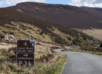 Eine einfache Straße. Daneben links ein Doppelschild. Darauf steht: Bealach Mileata MILITARY ROAD und darunter ein Pfeil nach rechts und GLENMACNASS Parkplatz und Aussichtspunkt