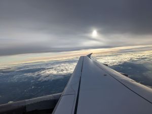 Flügelspitze eines Flugzeugs. Unten sind Wolken zu sehen, und über der Spitze ebenfalls eine Wolkendecke, in der die Sonne zu erkennen ist.