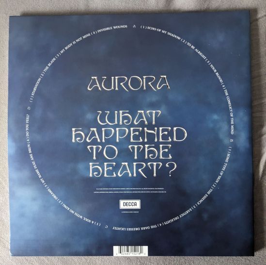 Rückseite meiner AURORA-Albumausgabe. In der Mitte steht in Großbuchstaben AURORA WHAT HAPPEND TO THE HEART? Im Kreis um diese zentrale Textgestaltung sind im Kreis alle Lieder, und auf welcher Seite sie sind, aufgeführt. Der Hintergrund ist blau wie der Himmel mit Wolken, in unterschiedlichen Blauabstufungen, bis Schwarz.