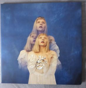 Vorderseite des Albums von AURORA. Aurora ist dreifach zu sehen, das silberne Herz vor sich, gedankenvoll nach oben blickend. Drt Hintergrund ist blau wie der Himmel mit Wolken, in unterschiedlichen Blauabstufungen, bis Schwarz.