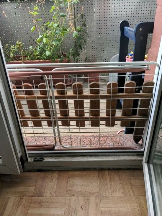 Geöffnete Balkontür, mit Blick zum Balkon hinaus. Vor der Tür liegt ein Wäscheständer quer davor, und vor dem Wäscheständer steht noch ein Holzzaunteil.