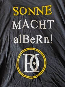 Schrift auf schwarzem Grund; SONNE MACHT alBeRn! Darunter die stilisierten Initialen vom Final Destination Club