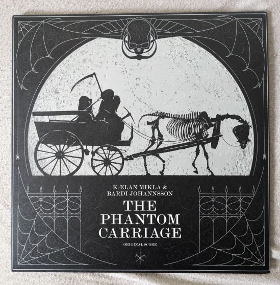 Cover der LP von Kælan Mikla & Bardi Johannsson THE PHANTOM CARRIAGE Orchestral Score Das Cover ist schwarz-weiß. Im Zentrum ist eine einfache Kutsche, das von einem Skelettpferd gezogen wird. Auf dem Wagen sitzen drei verhüllte Gestalten. Diese haben eine Sense, eine Spitzhacke und ein Schwert dabei. Über ihnen im Zentrum schwebt ein Totenkopf mit Fledermausflügeln. Unten in den Ecken sind Spinnennetze und ein Zaun links und rechts. An beiden Seiten lassen sich kleine Spinnen an einem Faden herab.