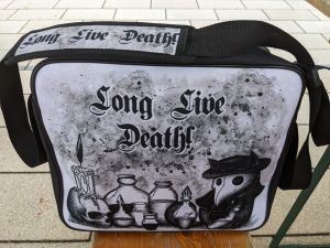 Umhängetasche mit der Beschriftung "Long Live Death!" Eine kleine Figur, wahrscheinlich Katze, mit Hut und Pestmaske sitzt rechts von mehreren kleinen Flaschen und Falkons. Ganz links ist ein Totenschädel mit brennender Kerze darauf.